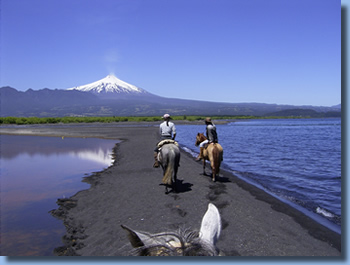 Two riders on horseback riding on a beach in front of Villarrica volcano